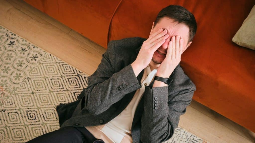 A man sitting on the floor leaning against a sofa while covering his face with both hands.