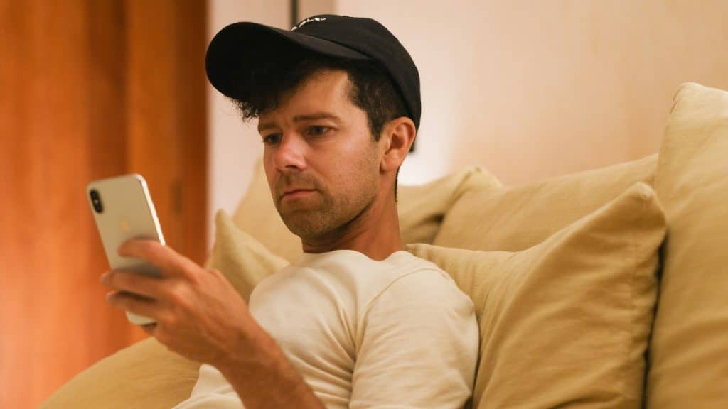 A man wearing a black baseball cap looks down at a smartphone in his hand.