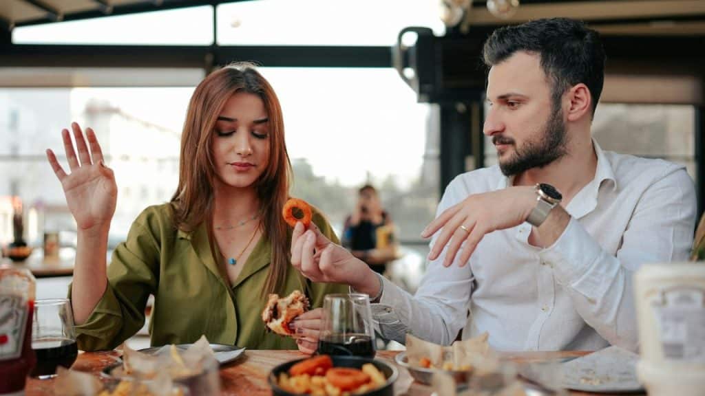 A man at a restaurant table holds an onion ring toward a woman with an upturned hand.