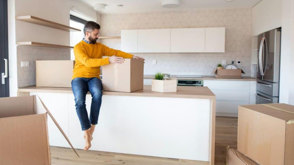 A man in a yellow sweater sits on a kitchen counter near cardboard boxes.