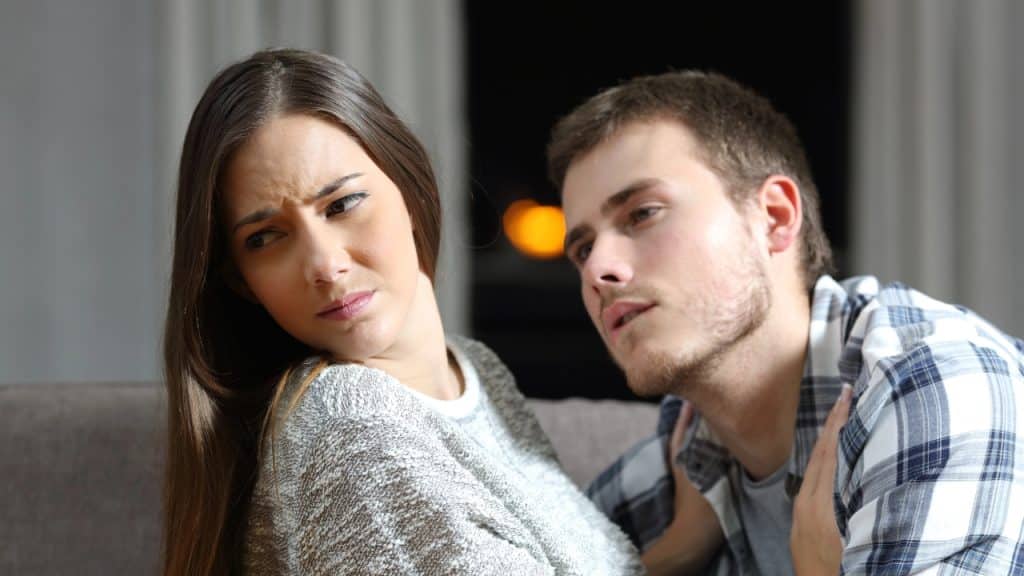 A woman with a frowning expression looks away while a man leans toward her shoulder.