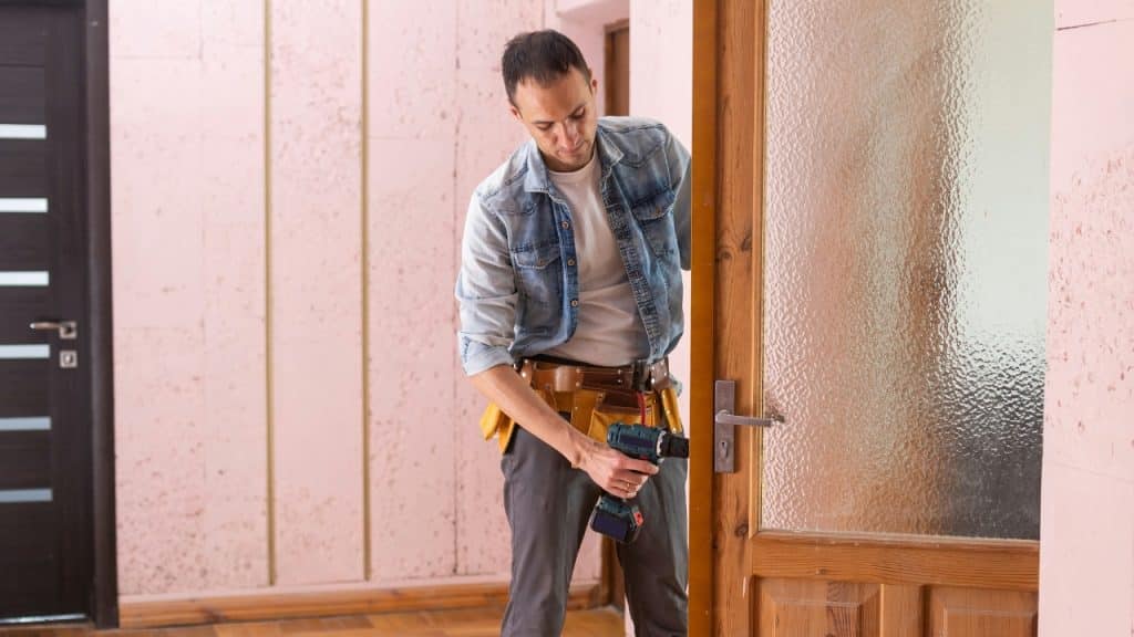 A man wearing a tool belt uses a power drill to repair a wooden door.