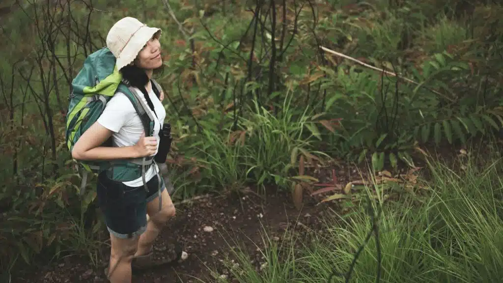 A woman wearing a sun hat and a green backpack hikes through a grassy area.