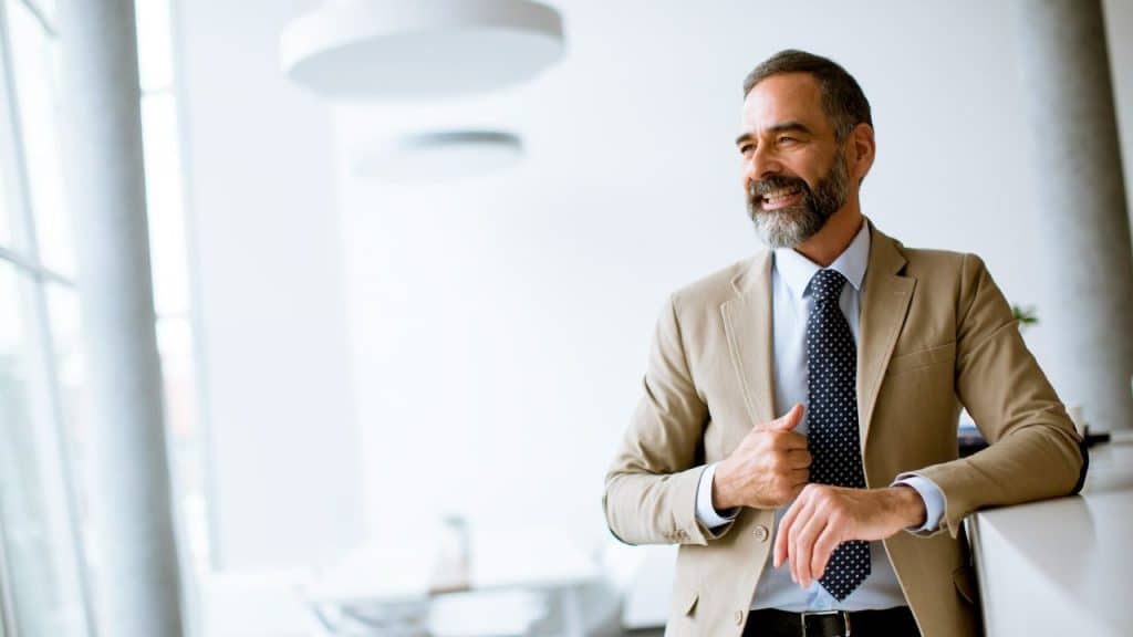 A smiling man with a beard wearing a tan suit leans against a white counter.