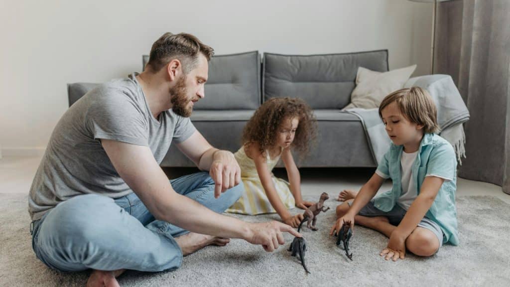 A man and two children sit on a rug playing with small plastic dinosaur figures.