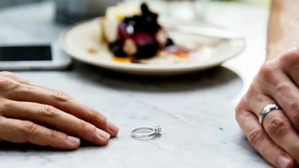 A silver diamond ring sits on a marble table next to hands and a plate of food.