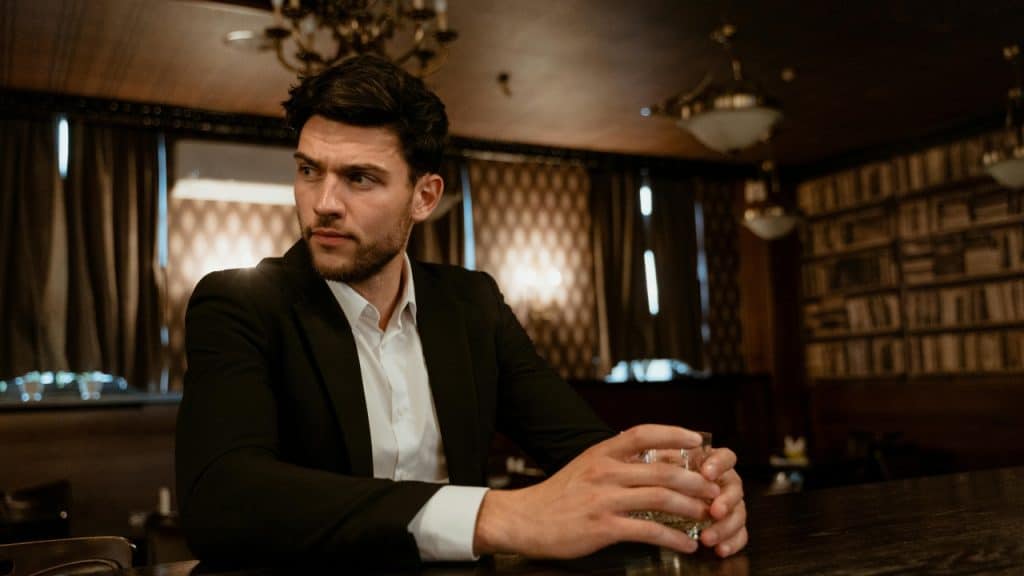 A man in a black suit sits at a bar holding a glass and looking away.