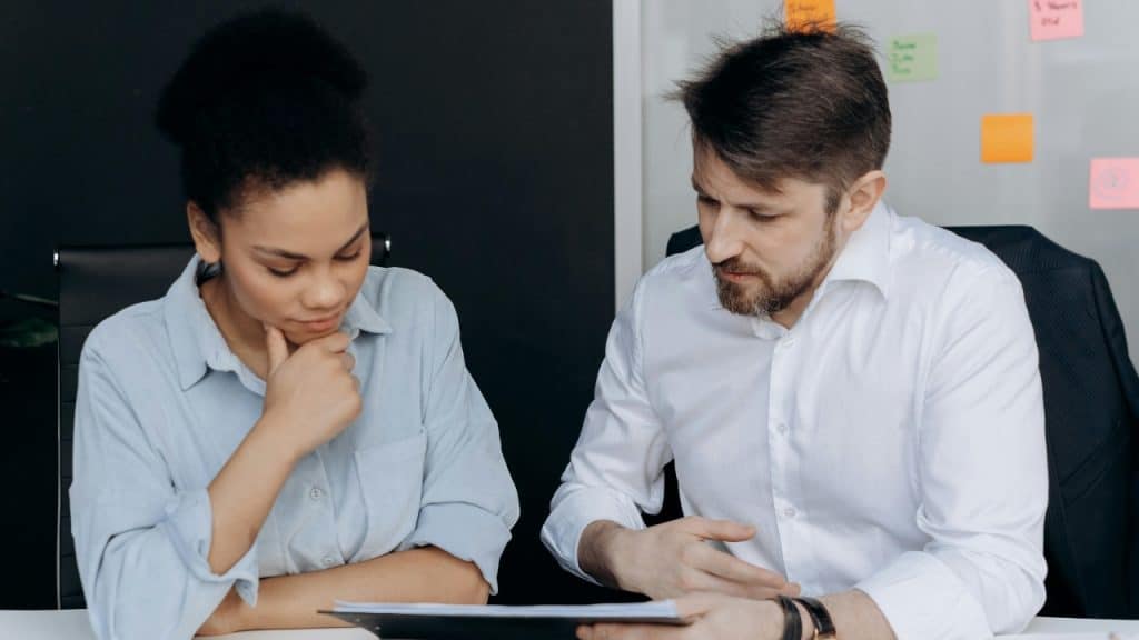 A man and woman sit at a white table, focusing on a black clipboard document.