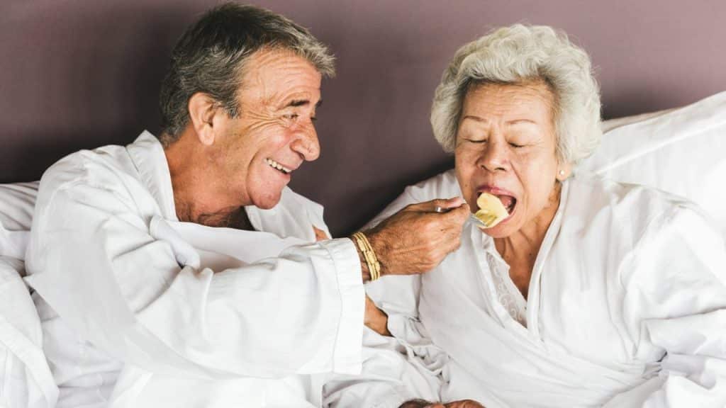 A smiling man in white clothing feeds a piece of fruit to a seated woman.