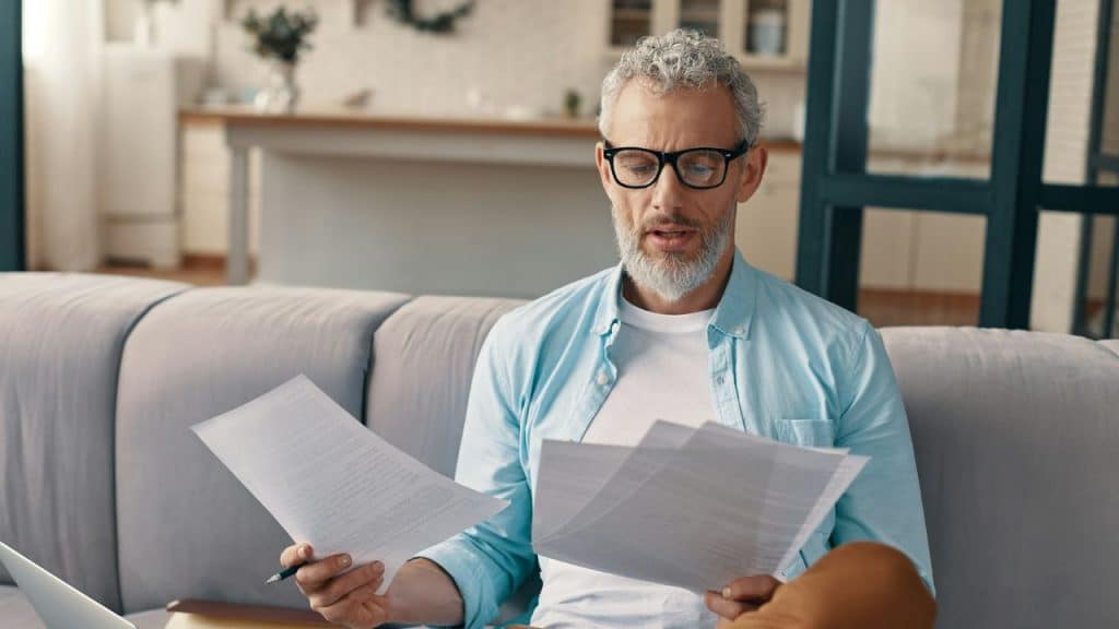 A man with glasses and a gray beard reviews several paper documents on a sofa.