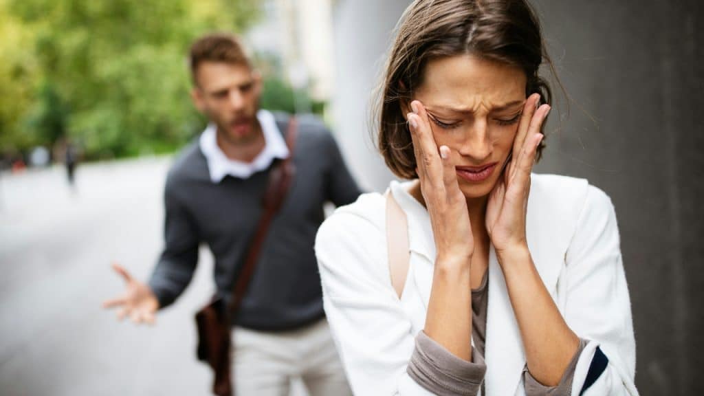 A woman holds her head with both hands while a man gestures behind her outdoors.