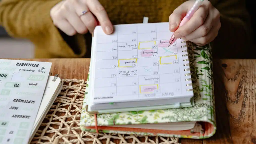 Close-up of hands using a pink pen to write in a spiral-bound monthly planner notebook.