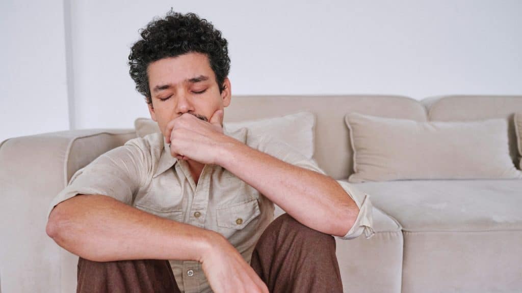 A man with curly hair sits on the floor with his hand over his mouth.