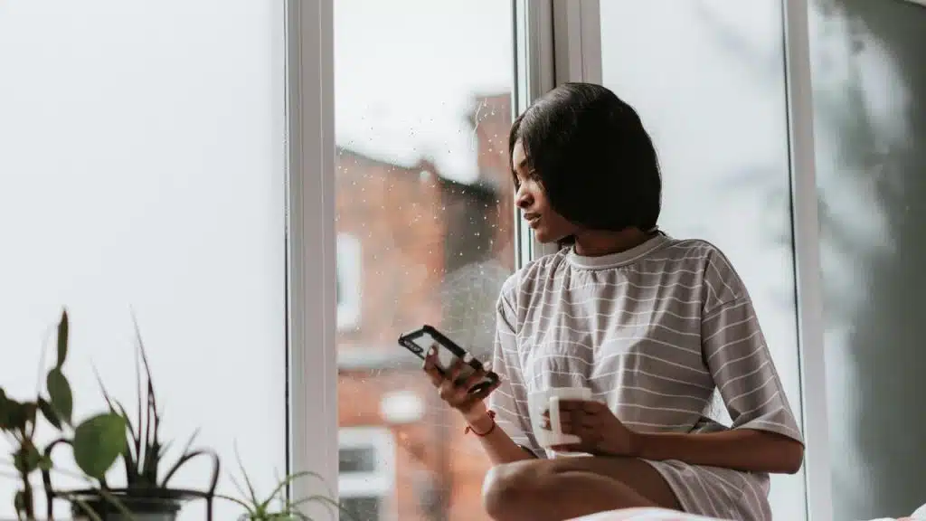 A woman sitting by a window holding a phone and a mug.