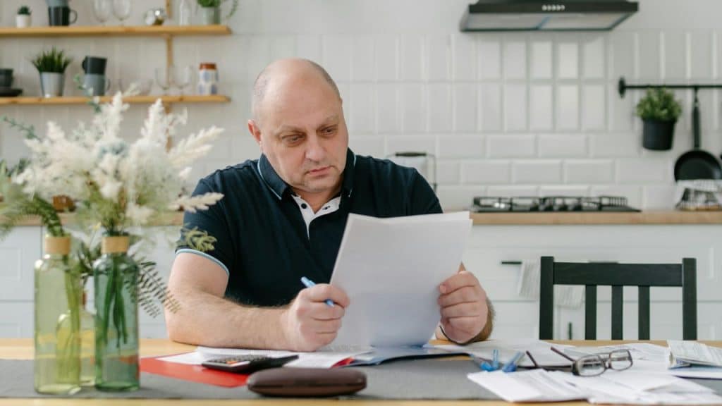 A man sits at a kitchen table holding a white paper with documents spread out.