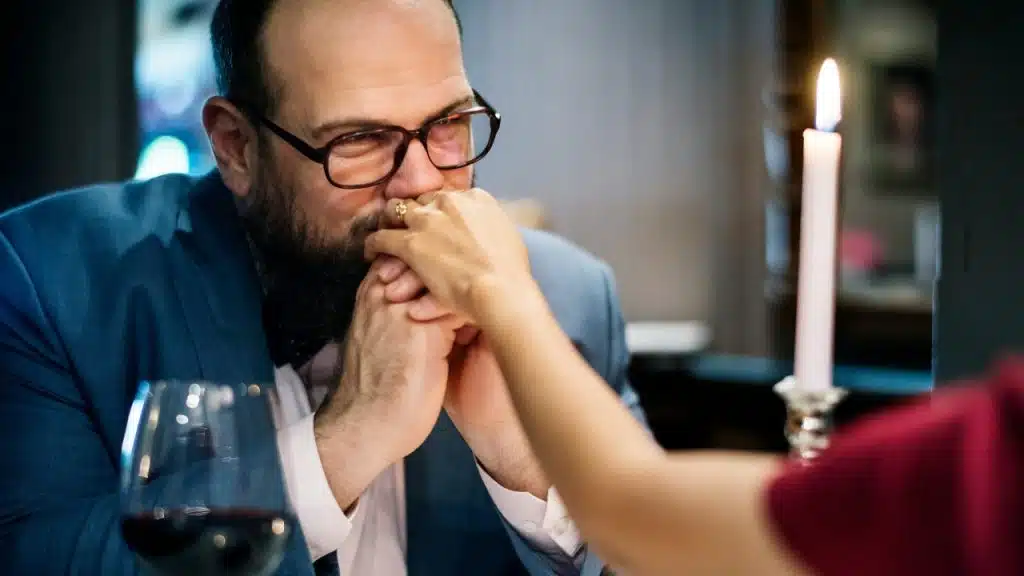 A man with glasses and a beard holds someone's hand near a lit white candle.