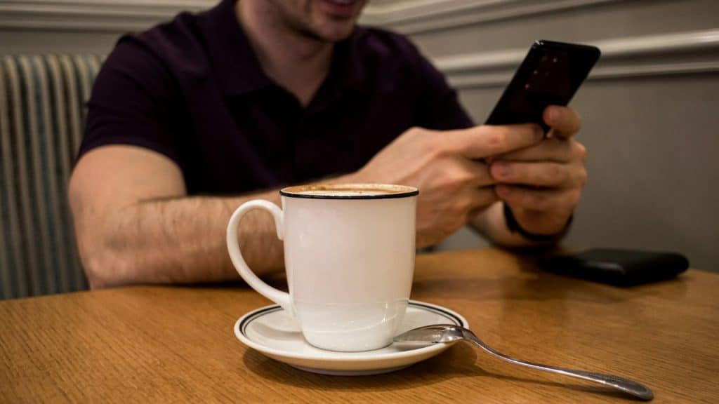 A man in a dark shirt uses a smartphone behind a white coffee mug on a table.