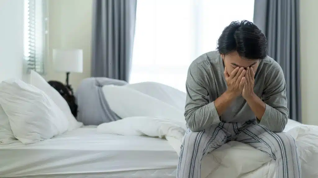 A man covering his face with his hands is sitting on one side of the bed, while his wife is sitting on the other side of the bed.