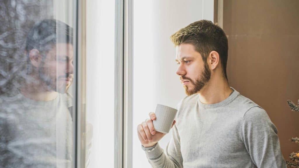 A man holding a mug while looking thoughtfully out a window.