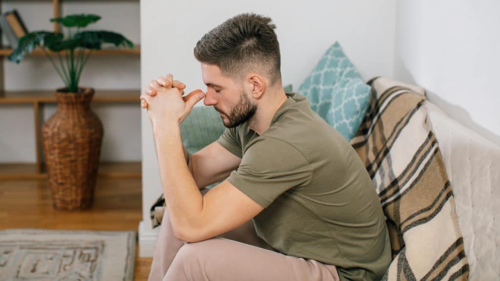 A man in an olive shirt sits on a sofa with his hands clasped and eyes closed.