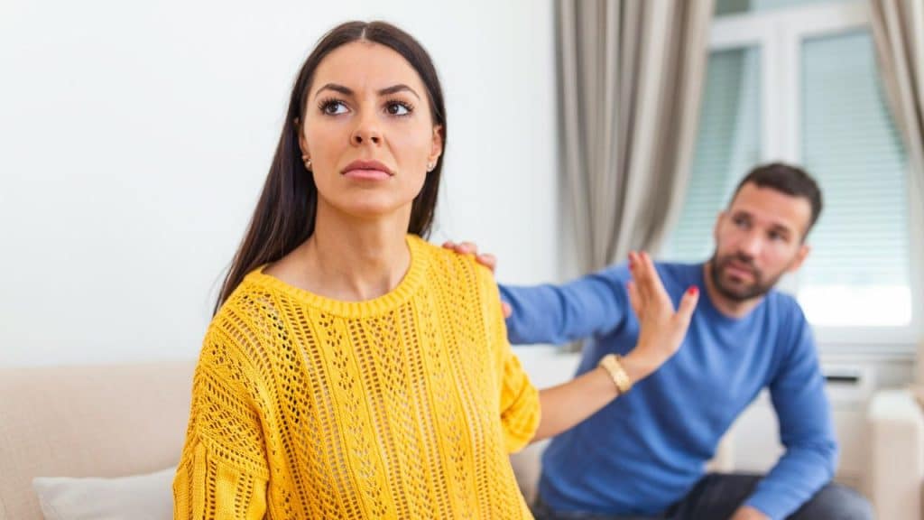 A woman in a yellow sweater looks away while a man reaches toward her shoulder.