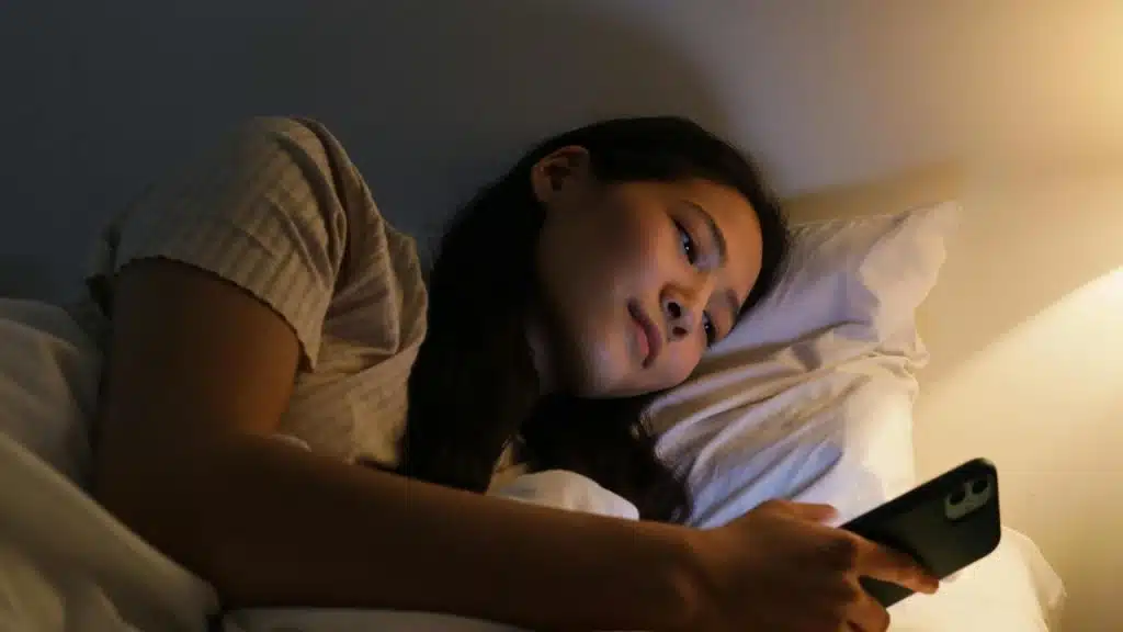 A woman lying in bed at night, looking at her phone.