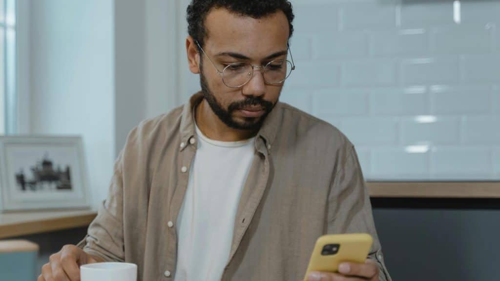 A man wearing glasses looking at his phone while holding a coffee cup.