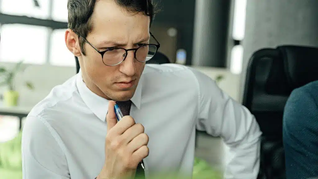 A man in glasses holding a pen and looking concerned.