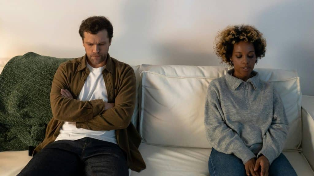 A man and woman sit apart on a white sofa with neutral expressions and folded arms.