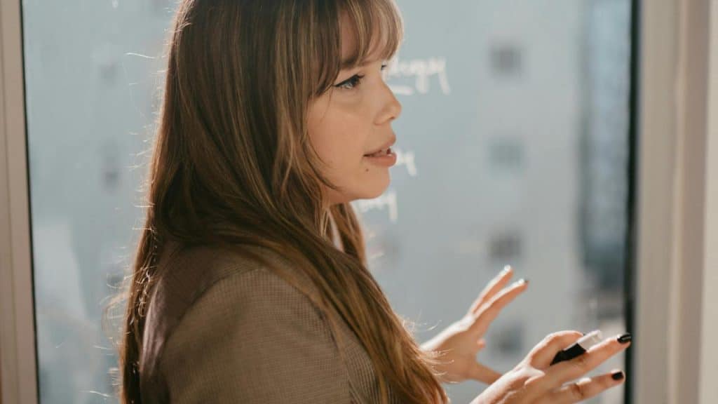 A woman in profile gesturing while speaking near a window.
