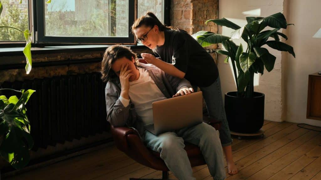 A woman leans over a man with a laptop while he covers his face with one hand.