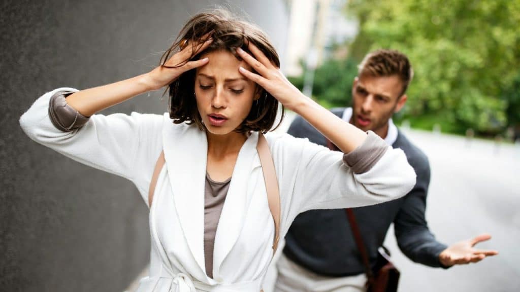 A woman holding her head looks away from a man who is gesturing behind her.