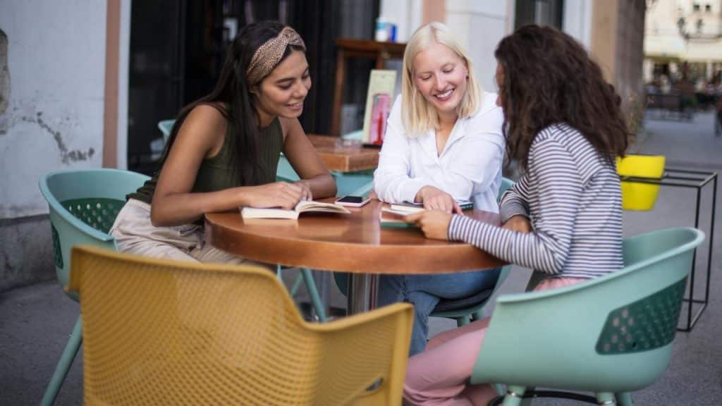A group of people sit at an outdoor table, smiling and looking at a book together.