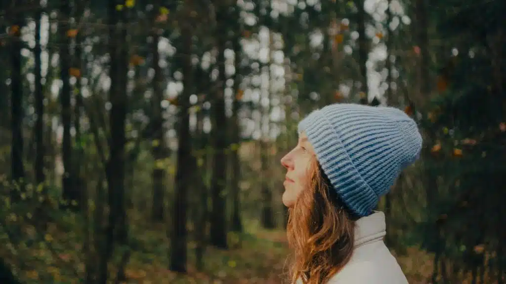 A woman in a knit hat looks upward while standing in a quiet forest.