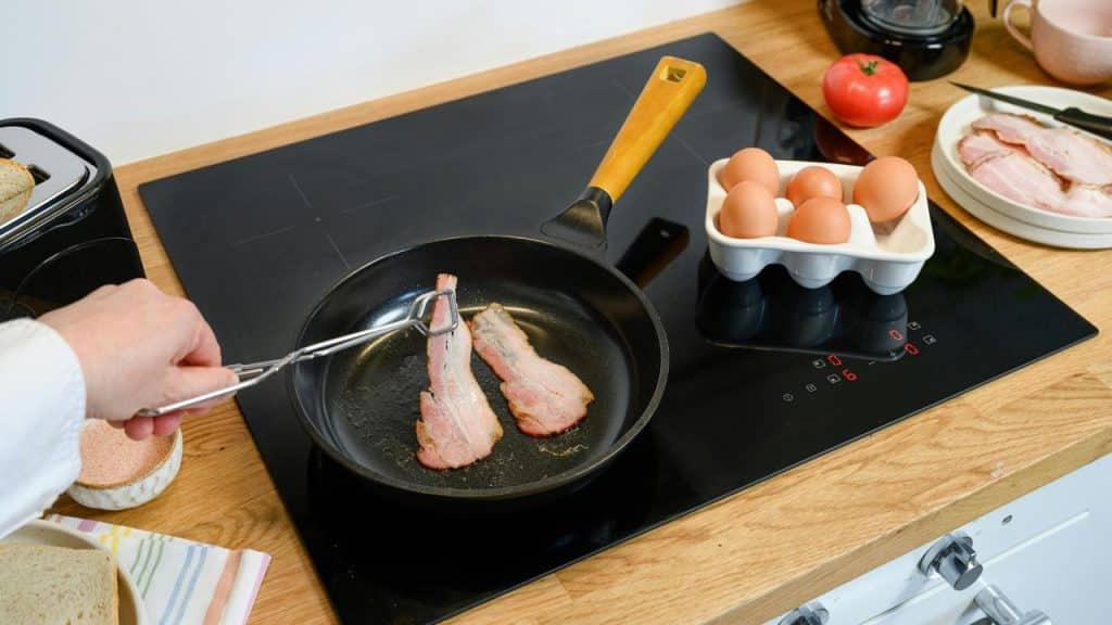 A hand flips bacon strips cooking in a frying pan on a stovetop.