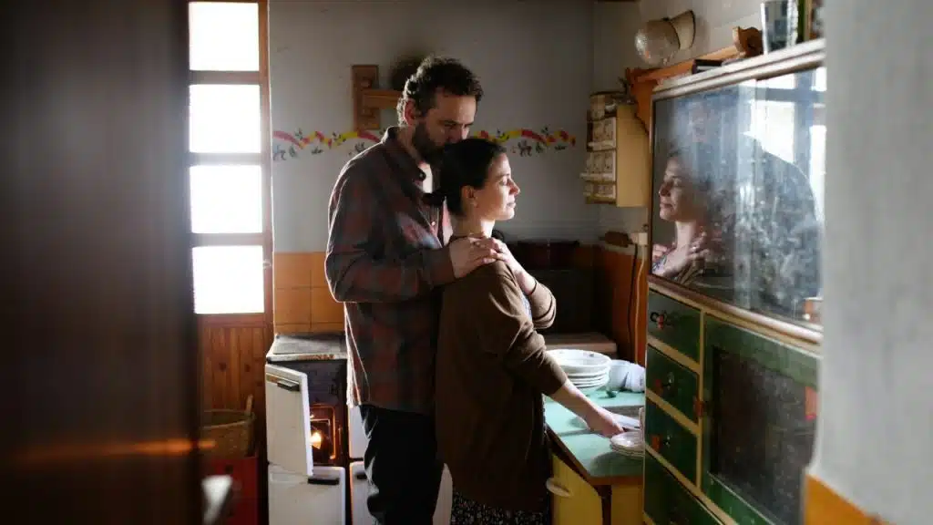 In a dimly lit kitchen, a man stands behind a woman with his hand on her shoulder.