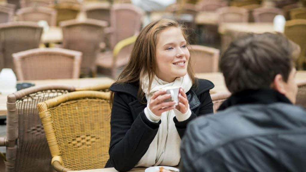 A woman in a black coat smiles while holding a mug at an outdoor cafe.