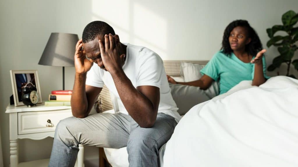 A man sits on a bed holding his head while a woman gestures behind him.