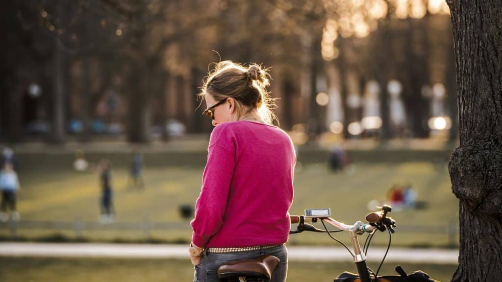 A woman standing beside a bicycle in a sunlit park.
