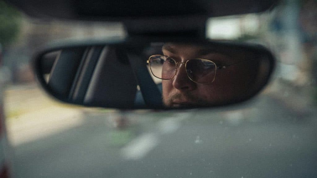 A man wearing glasses reflected in a car’s rearview mirror while driving.