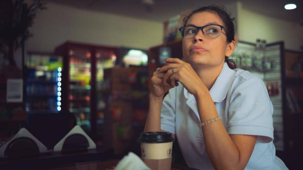 A woman wearing glasses, sitting in a café and looking thoughtfully to the side.