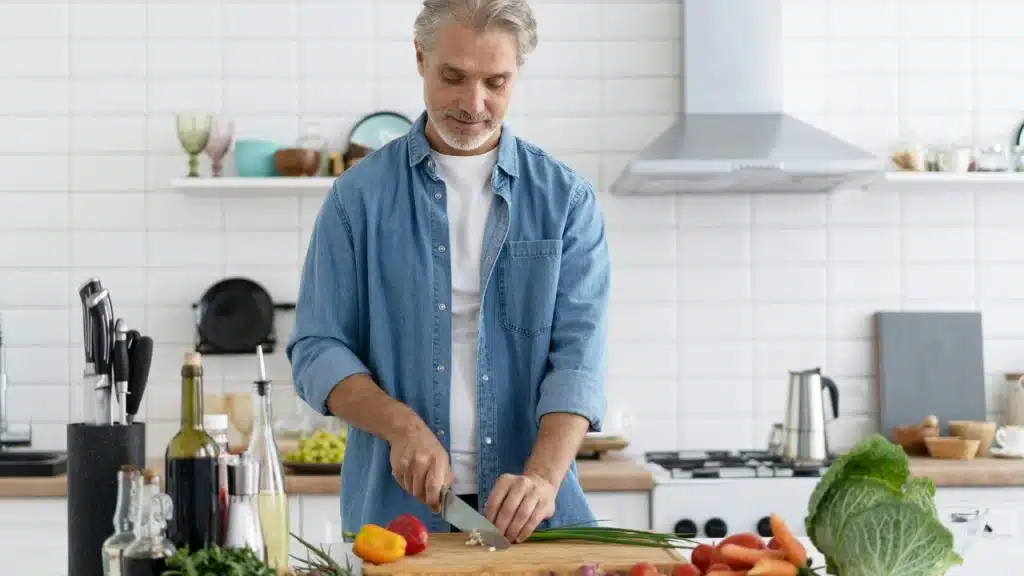 A man in a denim shirt chops green onions on a wooden board in a kitchen.