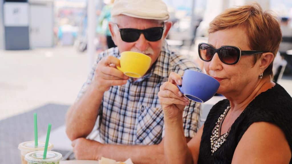 An older man and woman wearing sunglasses sit outdoors while drinking from colorful ceramic cups.