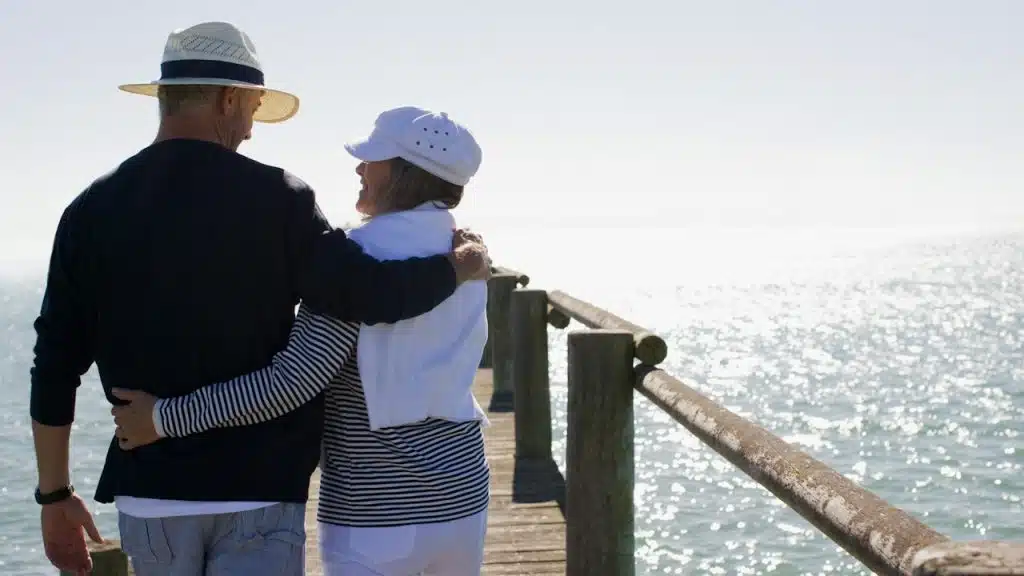 A couple walking down a pier with their arms around each other’s backs and smiling at each other.
