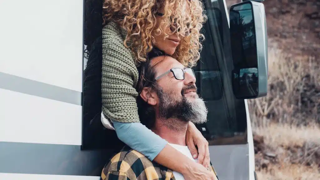 A woman with curly hair leans out of an RV window, wrapping her arms affectionately around a smiling man standing outside.