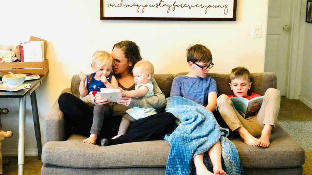 Mother sits on a sofa holding two toddlers while two older children sit nearby reading books at home.