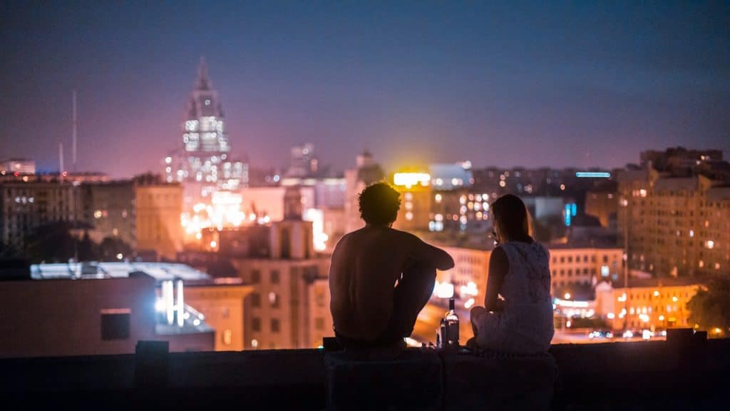 A couple sitting on a building overlooking an urban landscape at night.