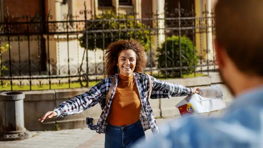A smiling woman opens her arms while holding a map on a sunny city street.