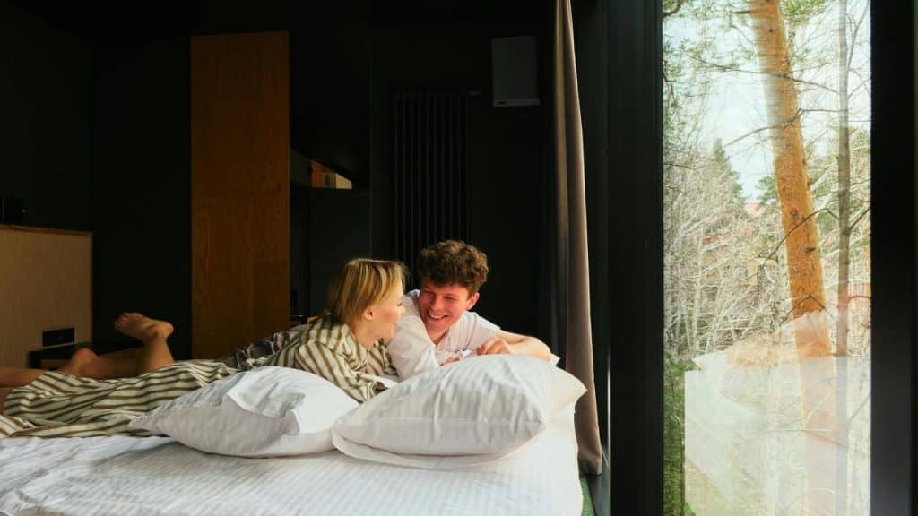 A couple laughs together on a bed beside a large window with a forest view.