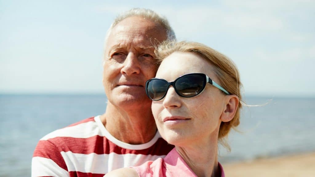 An aged man embracing his wife in sunglasses while both enjoy their rest by the seaside on a summer day.